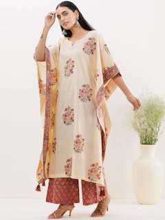 A person wearing a light cream-colored traditional outfit with floral patterns poses in front of a pale background. The outfit features a long tunic and wide-leg pants, accessorized with large earrings and brown heeled sandals. Next to the person is a tall white vase containing leafy green plants.