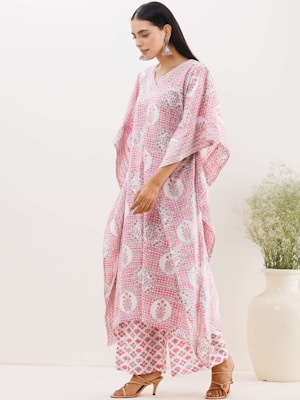 A woman in a light-colored room wearing a long, loose-fitting pink and white patterned dress and matching wide-legged pants. She has long dark hair, is wearing dangling earrings, and is standing in front of a light background next to a large, white vase with green foliage.