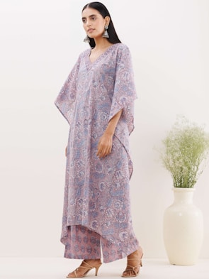 A woman is wearing a flowy, patterned, pastel-colored outfit with wide sleeves and matching pants. She has long, dark hair and is accessorized with large, dangling earrings. She is standing next to a tall white vase filled with delicate greenery, and she is wearing high-heeled, open-toed shoes.
