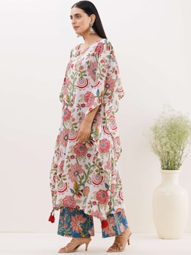A person stands wearing a floral-patterned dress with a combination of red, blue, and green colors. The outfit features intricate designs with leaf motifs. The person is accessorized with large earrings and heeled sandals. A white vase with green plant branches is visible in the background, set against a plain light-colored wall.