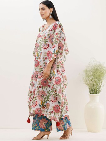 A person stands wearing a floral-patterned dress with a combination of red, blue, and green colors. The outfit features intricate designs with leaf motifs. The person is accessorized with large earrings and heeled sandals. A white vase with green plant branches is visible in the background, set against a plain light-colored wall.