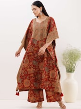 A woman is wearing a traditional red and orange patterned dress with intricate floral designs. Her outfit has wide sleeves and matching pants. She is accessorized with large dangling earrings and wearing heeled sandals. In the background, there is a light-colored vase with greenery against a plain wall.