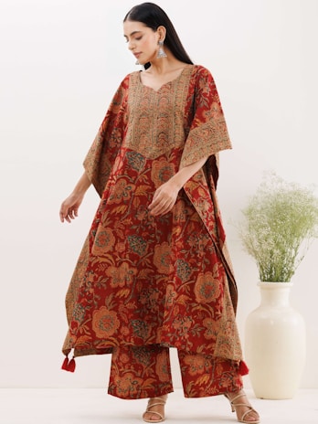 A woman is wearing a traditional red and orange patterned dress with intricate floral designs. Her outfit has wide sleeves and matching pants. She is accessorized with large dangling earrings and wearing heeled sandals. In the background, there is a light-colored vase with greenery against a plain wall.