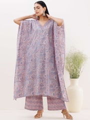 A woman is wearing a patterned, flowing dress with wide sleeves and matching pants. The garment features intricate floral designs in shades of purple and lavender. She is accessorized with large, dangling earrings and stands against a minimalistic background with a large white vase containing green foliage.
