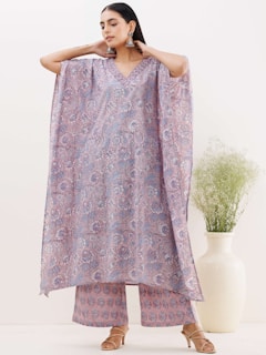 A woman is wearing a patterned, flowing dress with wide sleeves and matching pants. The garment features intricate floral designs in shades of purple and lavender. She is accessorized with large, dangling earrings and stands against a minimalistic background with a large white vase containing green foliage.