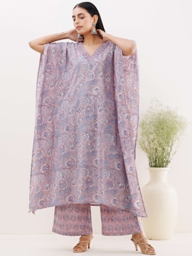 A woman is wearing a patterned, flowing dress with wide sleeves and matching pants. The garment features intricate floral designs in shades of purple and lavender. She is accessorized with large, dangling earrings and stands against a minimalistic background with a large white vase containing green foliage.