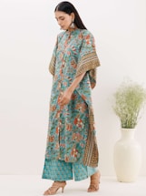 A woman is wearing a blue floral and patterned traditional outfit, consisting of a kurta and matching pants. She has long, straight hair and is adorned with large, intricate earrings. She is standing next to a tall white vase filled with green foliage.