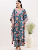 A woman is wearing a blue floral-patterned traditional outfit with wide sleeves and matching pants. The outfit is adorned with intricate designs in red and white. She stands beside a white vase containing green foliage. Her hair is tied back, and she is accessorizing with large earrings and sandals.