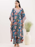 A woman is wearing a blue floral-patterned traditional outfit with wide sleeves and matching pants. The outfit is adorned with intricate designs in red and white. She stands beside a white vase containing green foliage. Her hair is tied back, and she is accessorizing with large earrings and sandals.
