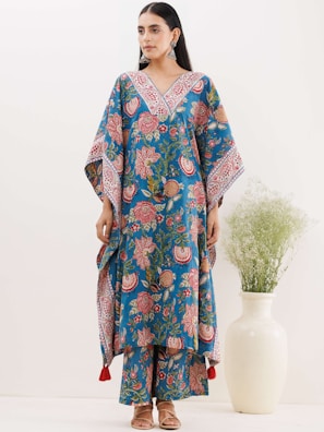 A woman is wearing a blue floral-patterned traditional outfit with wide sleeves and matching pants. The outfit is adorned with intricate designs in red and white. She stands beside a white vase containing green foliage. Her hair is tied back, and she is accessorizing with large earrings and sandals.