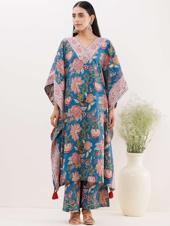 A woman is wearing a blue floral-patterned traditional outfit with wide sleeves and matching pants. The outfit is adorned with intricate designs in red and white. She stands beside a white vase containing green foliage. Her hair is tied back, and she is accessorizing with large earrings and sandals.