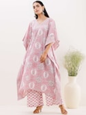 A woman wearing a loose-fitting, patterned garment in pink and white tones stands confidently. Her outfit features floral motifs and geometric patterns. She accessorizes with large, ornate earrings and brown heeled sandals. A tall, cream-colored vase with delicate greenery adds an elegant touch to the minimalist background.