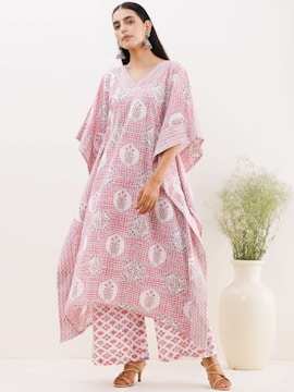 A woman wearing a loose-fitting, patterned garment in pink and white tones stands confidently. Her outfit features floral motifs and geometric patterns. She accessorizes with large, ornate earrings and brown heeled sandals. A tall, cream-colored vase with delicate greenery adds an elegant touch to the minimalist background.