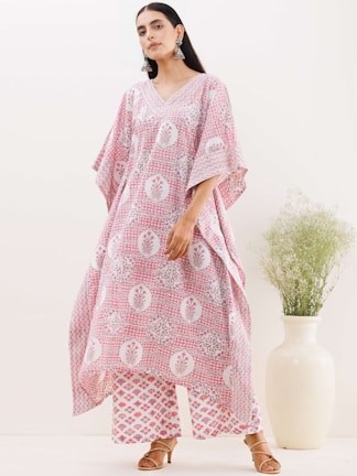 A woman wearing a loose-fitting, patterned garment in pink and white tones stands confidently. Her outfit features floral motifs and geometric patterns. She accessorizes with large, ornate earrings and brown heeled sandals. A tall, cream-colored vase with delicate greenery adds an elegant touch to the minimalist background.