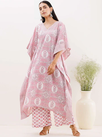 Confident plus-size woman in a flowing blush pink kaftan, smiling warmly against a soft beige background.