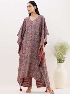 A person wearing a beautifully patterned traditional outfit with intricate designs, standing next to a large white vase containing greenery. The outfit features a mix of red, blue, and subtle floral or paisley patterns, with long, flowing sleeves and a layered hem. The individual is accessorized with statement earrings and sandals, adding an elegant touch to the overall attire.