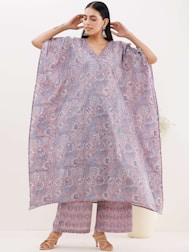 A woman is wearing an elegantly designed loose-fitting floral kaftan and matching pants. The garment features intricate blue and pink floral patterns. She accessorizes with large earrings and tan sandals, standing against a neutral background.