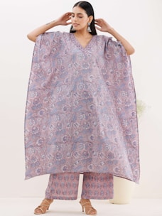 A woman is wearing an elegantly designed loose-fitting floral kaftan and matching pants. The garment features intricate blue and pink floral patterns. She accessorizes with large earrings and tan sandals, standing against a neutral background.