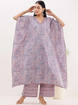 A woman is wearing an elegantly designed loose-fitting floral kaftan and matching pants. The garment features intricate blue and pink floral patterns. She accessorizes with large earrings and tan sandals, standing against a neutral background.