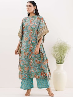 A person stands wearing a flowing, patterned garment in blue and orange with floral motifs. They have long dark hair and are accessorized with large earrings. The outfit includes a long top and matching pants. A large white vase with green foliage is placed nearby, adding a touch of natural elements.