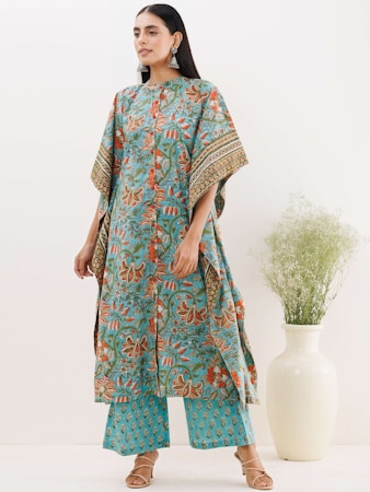 A person stands wearing a flowing, patterned garment in blue and orange with floral motifs. They have long dark hair and are accessorized with large earrings. The outfit includes a long top and matching pants. A large white vase with green foliage is placed nearby, adding a touch of natural elements.
