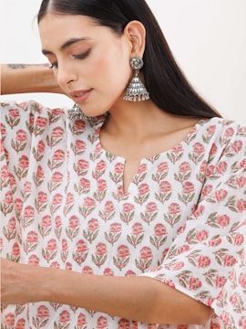 A woman with long dark hair wearing large, intricate silver earrings. She is dressed in a white garment featuring a floral pattern with pink and green hues.