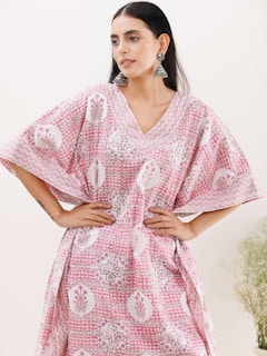 A woman with long dark hair wearing a loose-fitting, patterned dress with pink and white colors, and intricate floral designs. She has her hands on her hips and is accessorized with large, dangling earrings. The background is light with faint greenery visible on the right side.