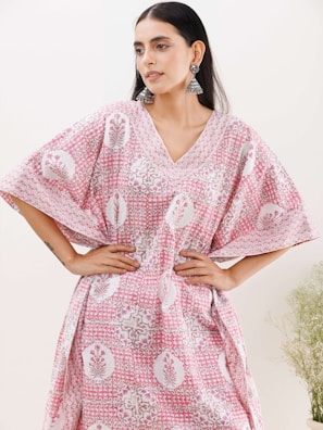 A woman with long dark hair wearing a loose-fitting, patterned dress with pink and white colors, and intricate floral designs. She has her hands on her hips and is accessorized with large, dangling earrings. The background is light with faint greenery visible on the right side.
