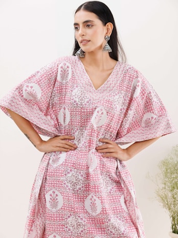 A woman with long dark hair wearing a loose-fitting, patterned dress with pink and white colors, and intricate floral designs. She has her hands on her hips and is accessorized with large, dangling earrings. The background is light with faint greenery visible on the right side.