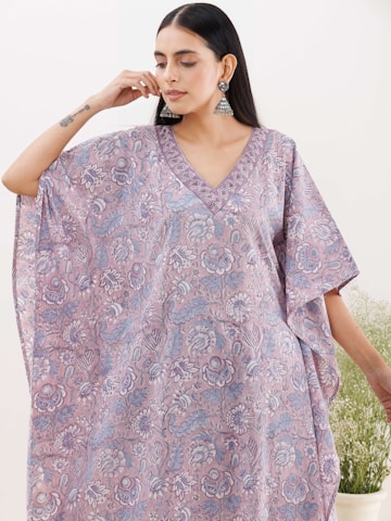 A woman is wearing a flowing kaftan with an intricate floral pattern in soft pastel colors. Her hair is straight and falls down her back, and she is adorned with large distinctive earrings. There is a subtle, serene expression on her face, and a tattoo is visible on her forearm. The background is minimalist, with some greenery.