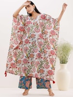 A person stands wearing a colorful floral-patterned outfit, featuring a loose-fitting top and wide pants. The top displays a variety of intricate floral designs in shades of red, pink, green, and blue. The person is accessorized with large earrings. Nearby, there is a white vase containing green foliage.