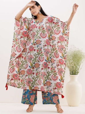 A person stands wearing a colorful floral-patterned outfit, featuring a loose-fitting top and wide pants. The top displays a variety of intricate floral designs in shades of red, pink, green, and blue. The person is accessorized with large earrings. Nearby, there is a white vase containing green foliage.