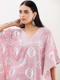 A person wearing a patterned pink and white garment, adorned with intricate floral designs. They have long, dark hair and are wearing large, ornate earrings. The mood is calm and stylish.