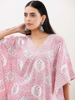 A person wearing a patterned pink and white garment, adorned with intricate floral designs. They have long, dark hair and are wearing large, ornate earrings. The mood is calm and stylish.