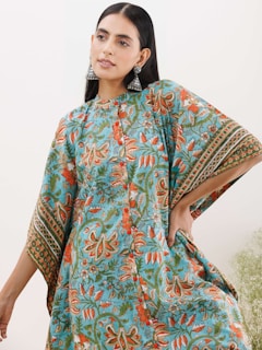 A woman is wearing a colorful, floral-patterned dress with orange, green, and blue hues. She accessorizes with large, ornate silver earrings and has long, dark hair parted in the middle. Her pose is relaxed and confident with one hand on her hip against a soft, neutral background.