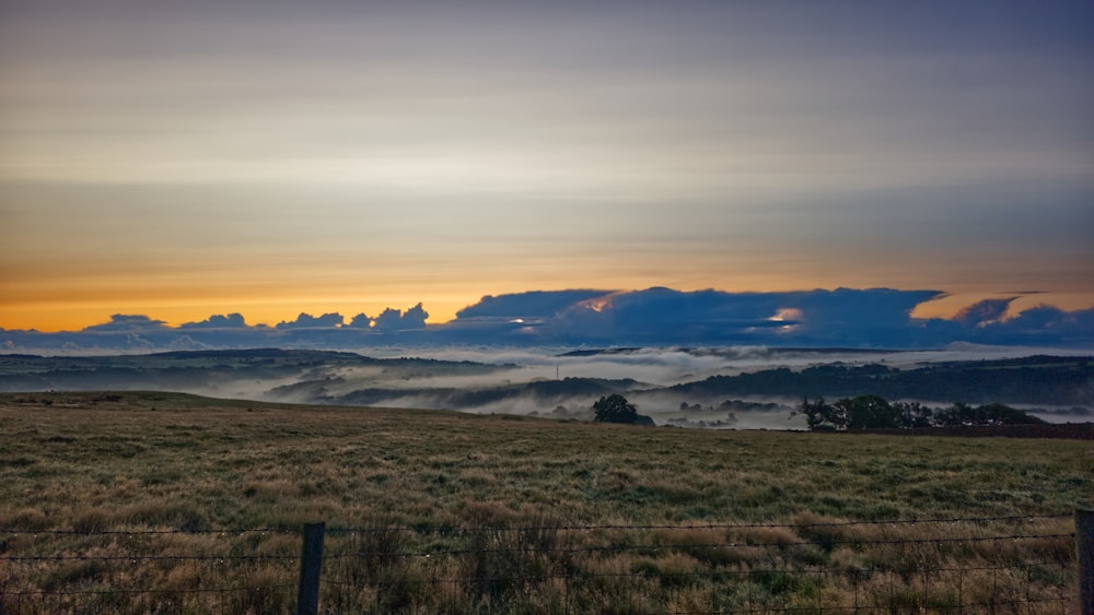 a field with a fence in the foreground and clouds in the background