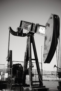 An oil pumpjack with a metallic, industrial structure is set against a clear sky, partially enclosed by a wire fence. In the background, another pumpjack can be seen, along with a barren landscape.
