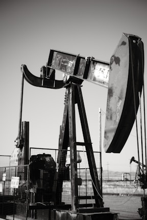 An oil pumpjack with a metallic, industrial structure is set against a clear sky, partially enclosed by a wire fence. In the background, another pumpjack can be seen, along with a barren landscape.