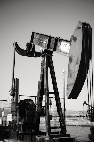 An oil pumpjack with a metallic, industrial structure is set against a clear sky, partially enclosed by a wire fence. In the background, another pumpjack can be seen, along with a barren landscape.