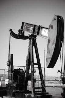 An oil pumpjack with a metallic, industrial structure is set against a clear sky, partially enclosed by a wire fence. In the background, another pumpjack can be seen, along with a barren landscape.