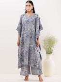 A person is wearing a stylish, loose-fitting outfit with a floral pattern in shades of blue and grey. The outfit comprises a long top with wide sleeves and matching wide-leg pants. The person is accessorized with large earrings and stands beside a tall, minimalistic white vase containing green foliage.