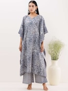 A person is wearing a stylish, loose-fitting outfit with a floral pattern in shades of blue and grey. The outfit comprises a long top with wide sleeves and matching wide-leg pants. The person is accessorized with large earrings and stands beside a tall, minimalistic white vase containing green foliage.