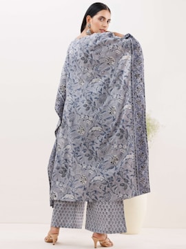 A person is elegantly dressed in a patterned shawl and matching trousers. The shawl displays intricate floral designs in shades of blue and grey. The trousers feature a smaller repetitive pattern. The person is wearing open-toed heels and large dangling earrings, standing against a plain, light-colored background.
