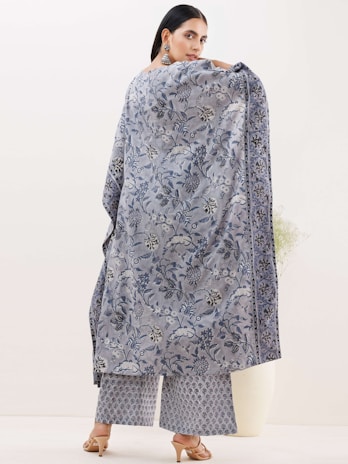 A person is elegantly dressed in a patterned shawl and matching trousers. The shawl displays intricate floral designs in shades of blue and grey. The trousers feature a smaller repetitive pattern. The person is wearing open-toed heels and large dangling earrings, standing against a plain, light-colored background.