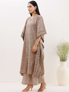 A woman stands against a plain light background wearing a floral-patterned outfit with wide sleeves and matching trousers. She accessorizes with large, traditional earrings and sandals. A white vase with green foliage is positioned to her right.