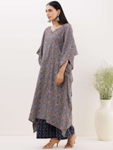 A woman is standing in a side pose, wearing a traditional kaftan with a geometric pattern in earthy tones. She has long hair and is accessorized with large dangling earrings. The background is plain with a large ceramic vase containing some greenery.