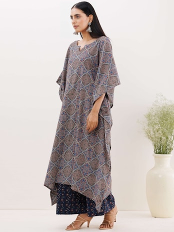 A woman is standing in a side pose, wearing a traditional kaftan with a geometric pattern in earthy tones. She has long hair and is accessorized with large dangling earrings. The background is plain with a large ceramic vase containing some greenery.