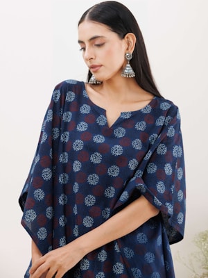 A person is wearing a loose-fitting outfit with a navy blue base color adorned with a repetitive pattern of red and white designs. The individual has long dark hair and is accessorized with large, ornate silver earrings. Their expression is calm and introspective.