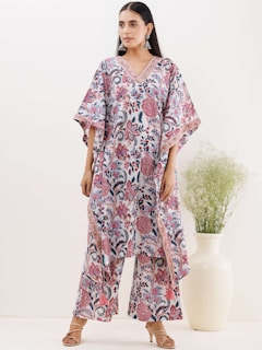 A woman is wearing a colorful floral outfit consisting of a V-neck top and matching wide-legged pants. The pattern includes pink and blue intricate floral designs. She is accessorized with large silver earrings and beige high heels. Standing beside a large white vase with green foliage.