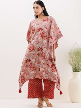 A person wearing a patterned, loose-fitting tunic with a floral design in red and beige tones. The outfit is accessorized with large, statement earrings and brown sandals. In the background, there is a white vase containing green foliage, set against a plain, light-colored wall.